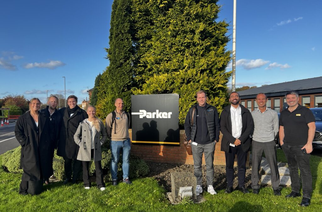Gruppfoto utomhus framför Parker Hannifins skylt med representanter från Colly Flowtech och Parker Hannifin under ett gemensamt besök. Solig dag med blå himmel, grönt gräs och leende deltagare.